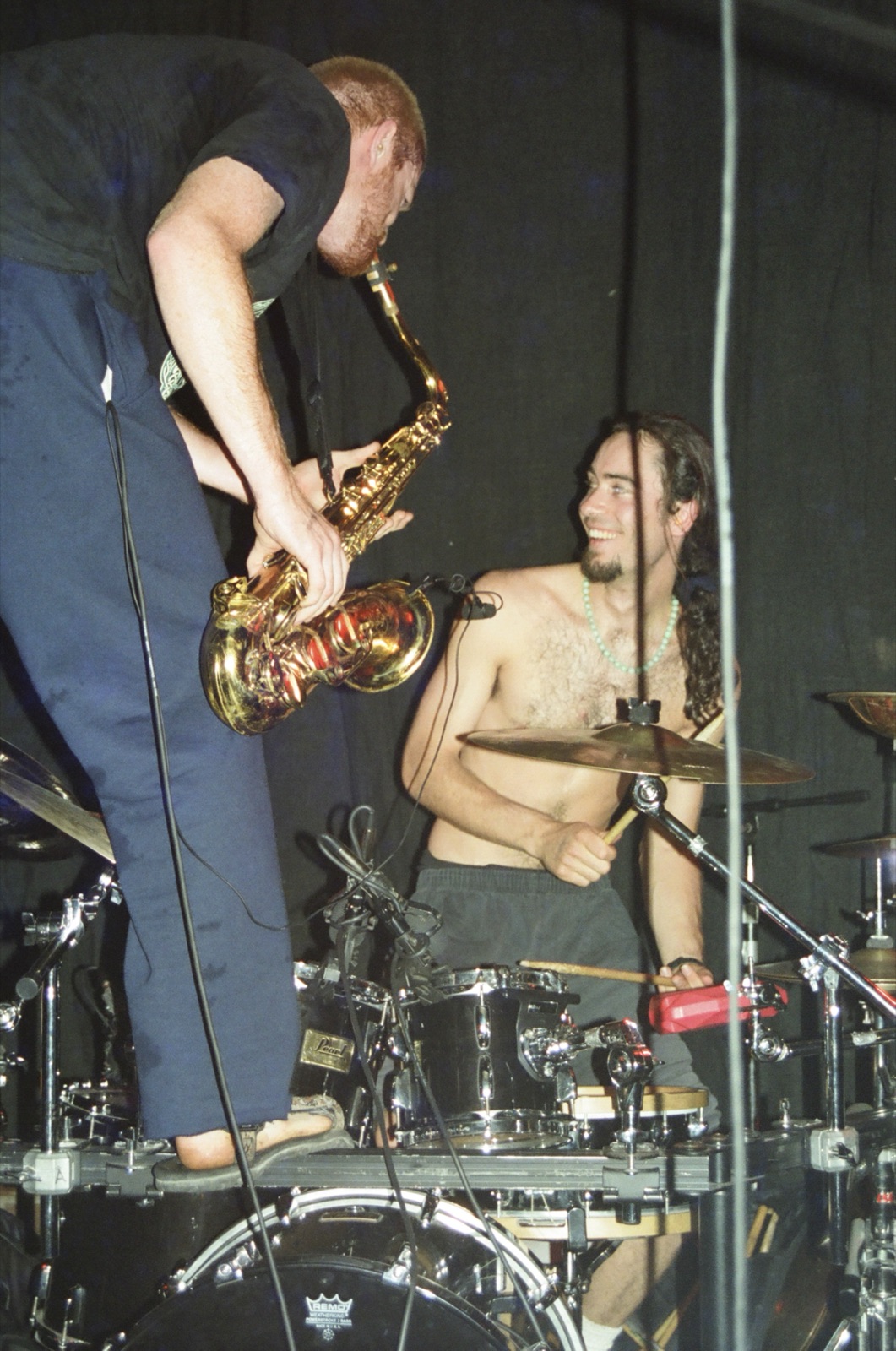 A saxophonist stands on the bass drum and leans over a grinning shirtless drummer mid-song.
